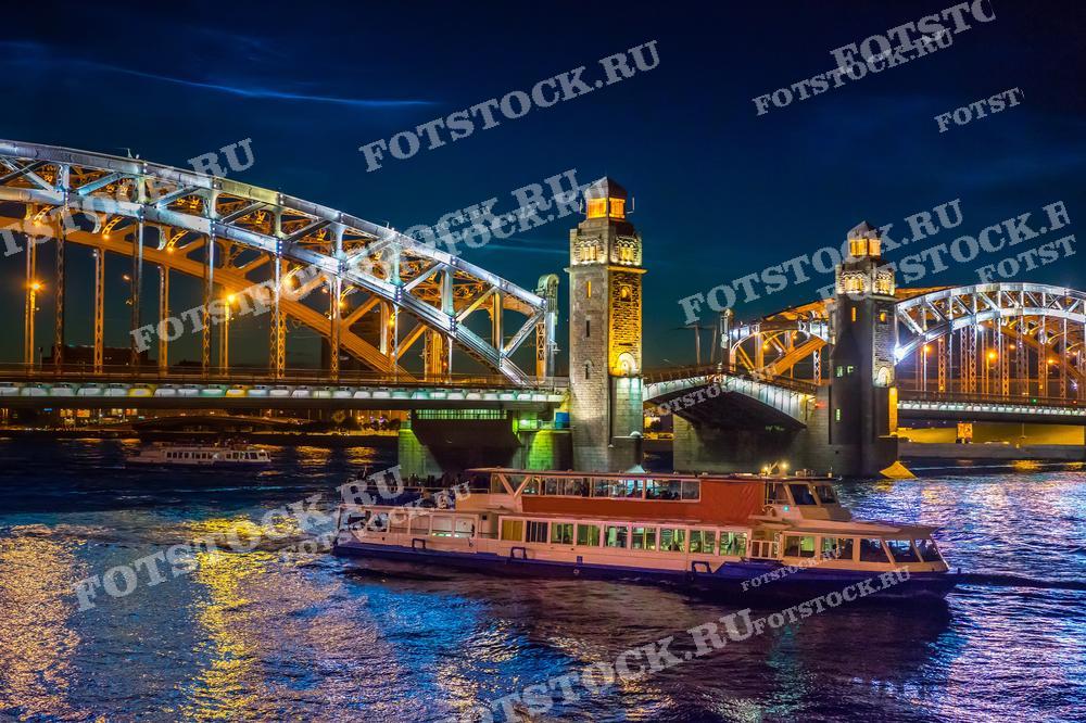 Мост и его архитектурные элементы выглядят великолепно на фоне ночного города, а на переднем плане виден туристический кораблик, плавающий по реке Нева. Это изображение подойдёт для использования в туристических публикациях и рекламных материалах, подчёркивающих красоту городской ночной жизни.